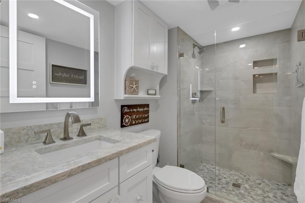 Contemporary bathroom featuring an illuminated mirror, a vanity with a light-toned countertop and integrated sink, brushed nickel faucet, a glass-enclosed shower with tiled walls, and a pebble-tiled shower floor