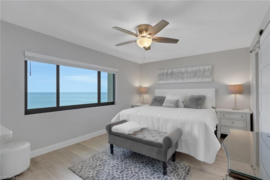 Expansive picture window showcasing water scenery, light wood-finish flooring, ceiling fan with integrated lighting, and a white barn door