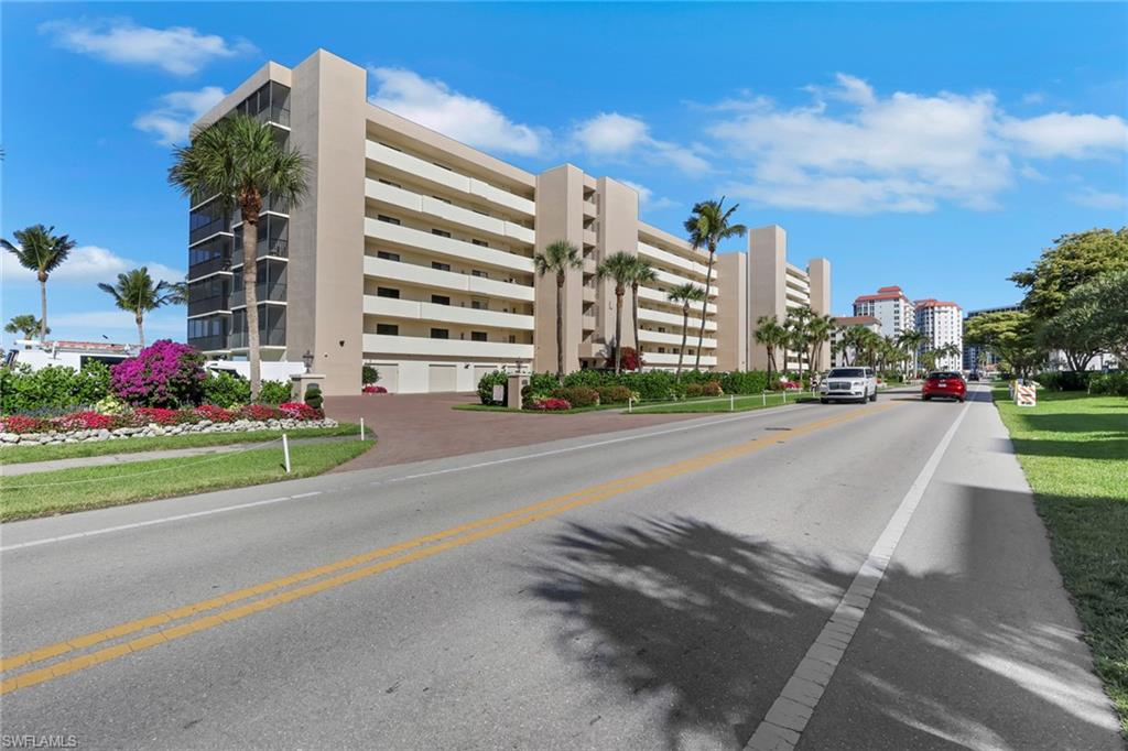 Multi-story building featuring extensive balconies, a red paver driveway, lush landscaping with vibrant flowers, and mature palm trees