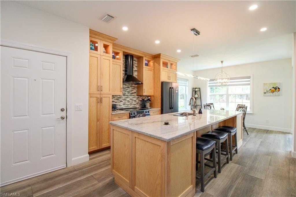 Kitchen with a center island with sink, pendant lighting, black refrigerator, backsplash, and wall chimney range hood