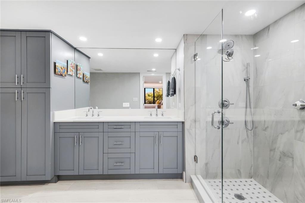 This bathroom features a spacious shower with a clear glass enclosure and marble-patterned tile, alongside a vanity with dual sinks, a large mirror, and ample gray cabinetry