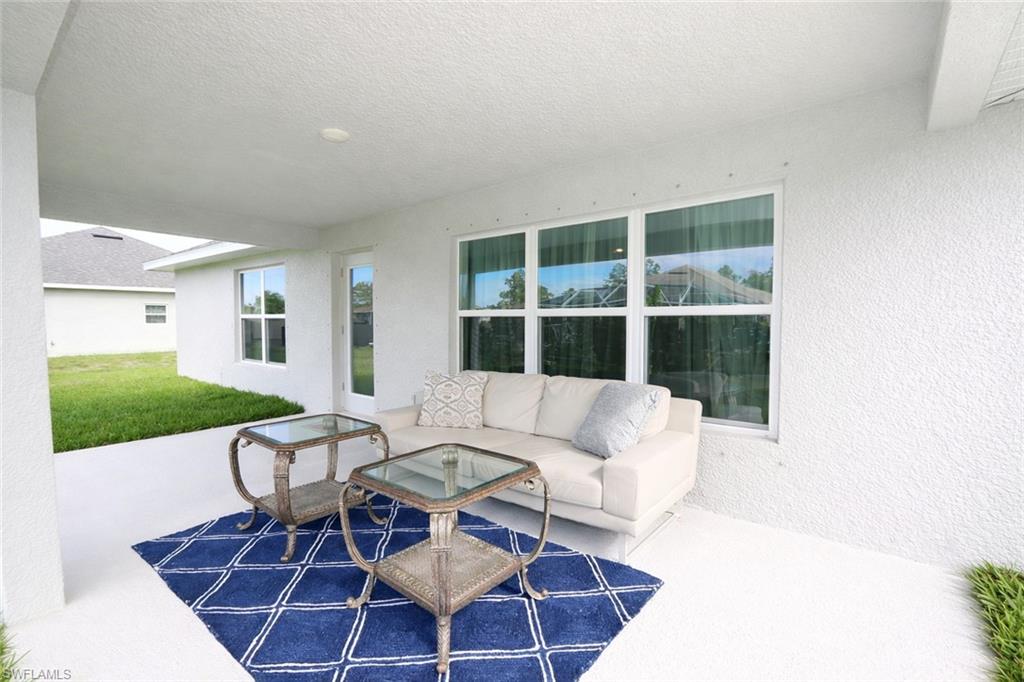 Covered outdoor patio area featuring a white textured wall, large windows, and a view of the surrounding green space