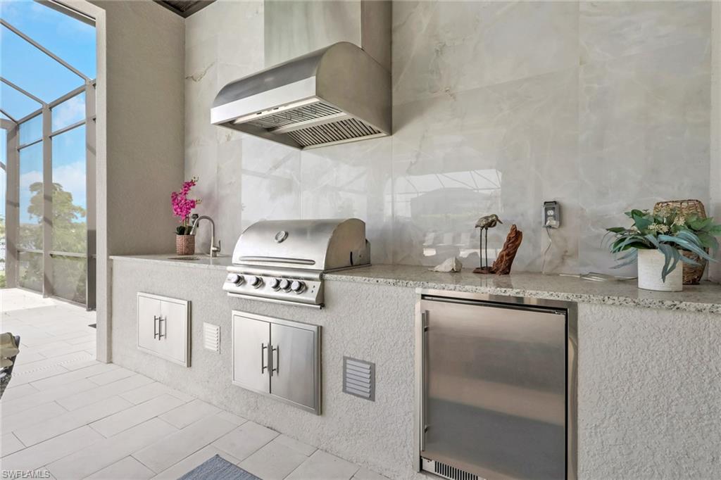 Outdoor kitchen featuring a built-in stainless steel grill, integrated range hood, under-counter refrigerator, and a sink with a stainless steel faucet