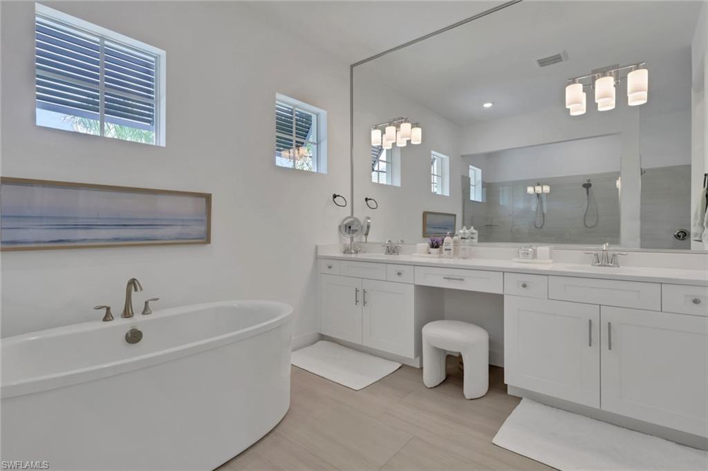 Spacious bathroom featuring a freestanding soaking tub with brushed nickel fixtures, an extended double vanity with white cabinetry and integrated sinks, a large mirror spanning the vanity, and light wood-finish flooring