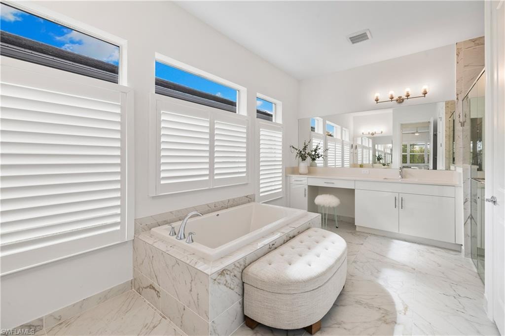 Spacious bathroom featuring a built-in soaking tub with tile surround, white plantation shutters, white vanity cabinetry, light-toned quartz countertops, and a glass-enclosed shower