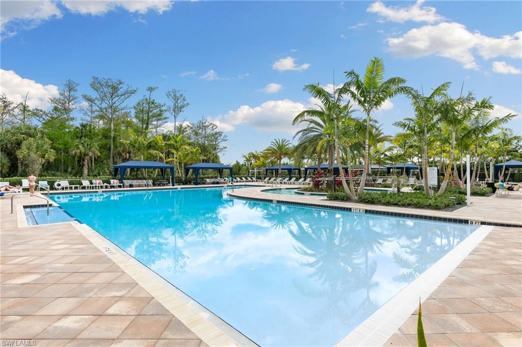 Expansive resort-style swimming pool featuring a large rectangular basin, surrounding paver patio, multiple poolside cabanas, and mature palm trees