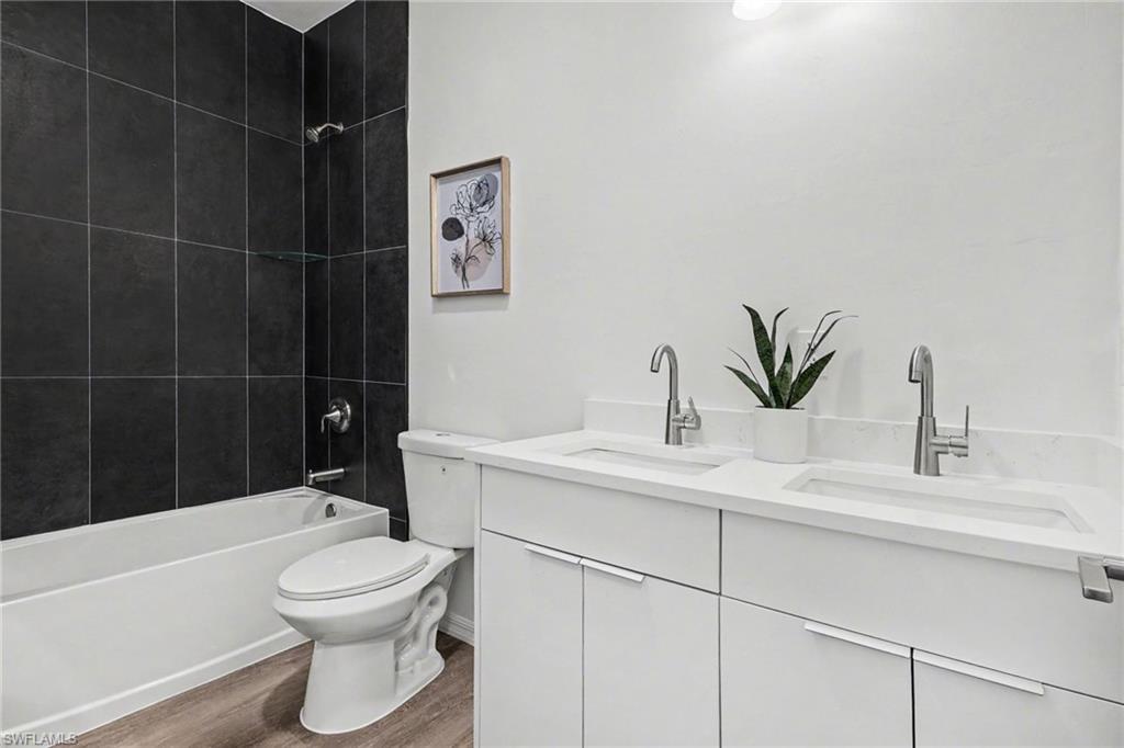 Bathroom featuring a white bathtub with dark tile surround, a dual vanity with undermount sinks, white countertops, and wood-finish flooring
