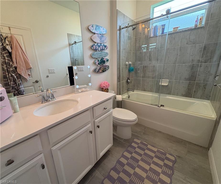 Bathroom featuring a single vanity with an integrated sink and light-toned countertop, white cabinetry, an elongated mirror, a toilet, a full-size bathtub with a sliding glass enclosure, and grey tiled flooring