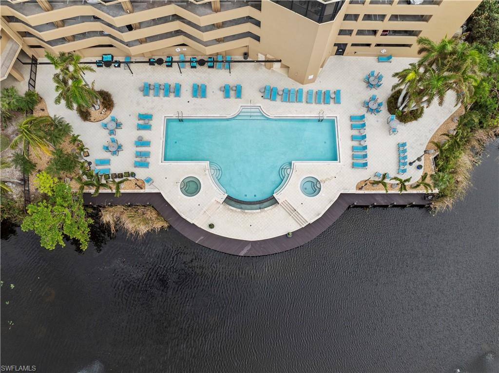 An aerial view highlighting the waterfront pool area, featuring a large swimming pool with integrated spas, a spacious deck with lounge chairs and tables, and mature palm trees