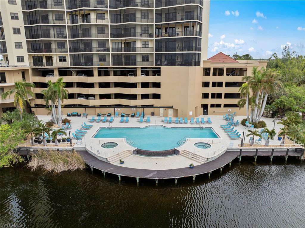 The property features a large outdoor pool and two circular hot tubs, surrounded by lounge chairs and palm trees, all set on a water-front deck