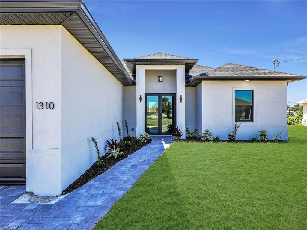 Contemporary stucco exterior with a covered entry, dark-framed double glass entry doors, a paver walkway, a manicured lawn, and professional landscaping  the color of the grass has been virtually enhanced