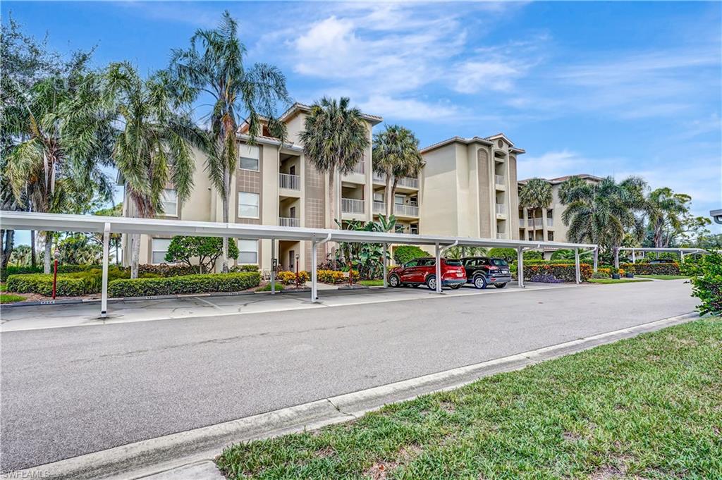 Multi-story building featuring covered parking structures, mature palm trees, landscaped shrubbery, and a paved parking area