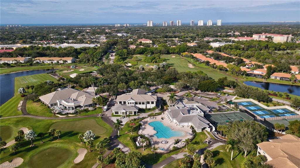 Expansive aerial perspective showcasing a golf course, multiple buildings with light-toned roofs, a large outdoor swimming pool, and several tennis courts
