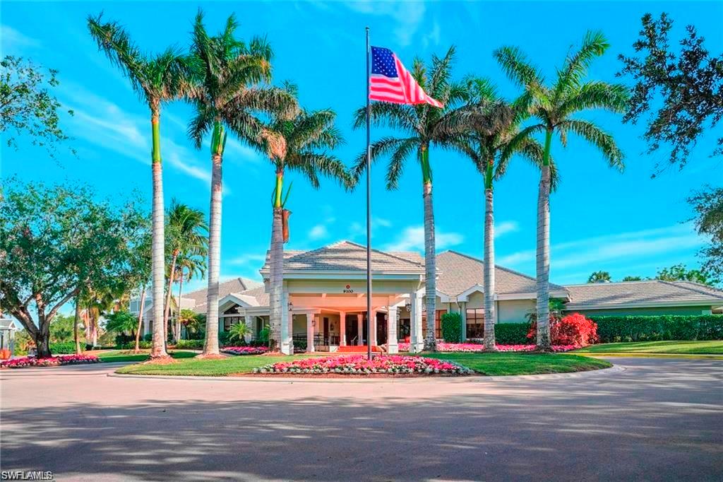Circular driveway entrance with a central landscaped island featuring a flagpole, bordered by palm trees and vibrant flowering plants