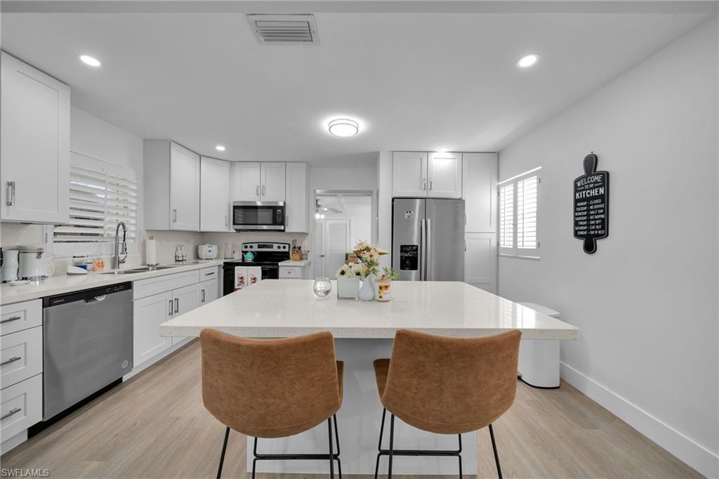 Contemporary kitchen featuring an expansive island with countertop seating, white shaker cabinetry, stainless steel appliances, and light wood-finish flooring