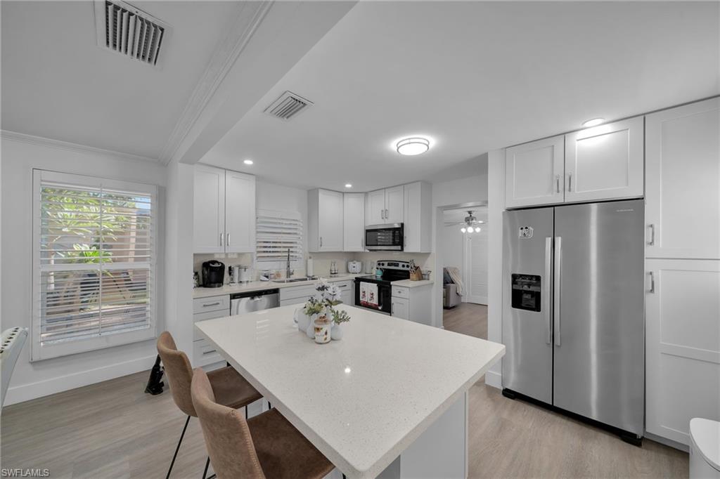 Contemporary kitchen featuring white shaker cabinetry, stainless steel appliances, a center island with countertop seating, a window with plantation shutters, and wood-finish flooring