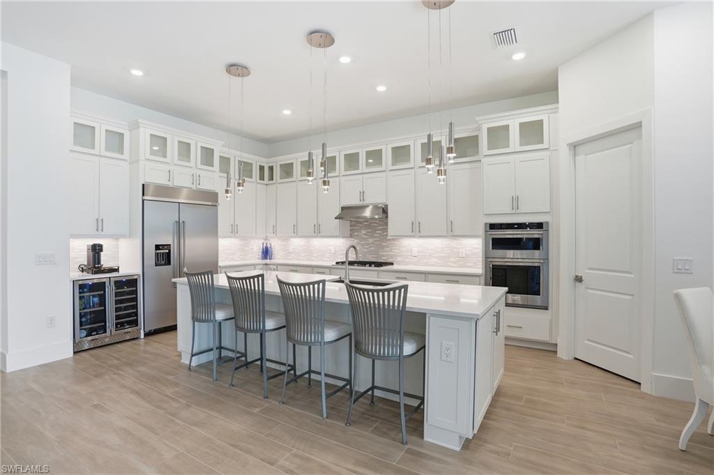 Spacious kitchen featuring an expansive island with seating, stainless steel appliances, white cabinetry with glass-front uppers, a tiled backsplash, and wood-finish flooring