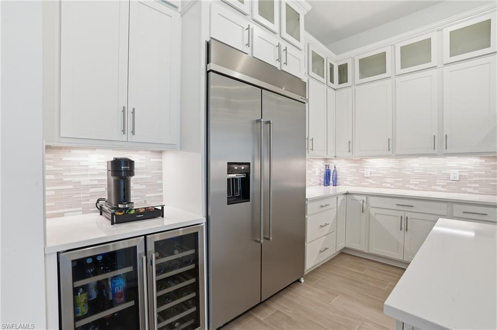 Gourmet kitchen with stainless steel refrigerator, white cabinetry, integrated beverage and wine coolers, light-toned tile backsplash, and light-colored countertops