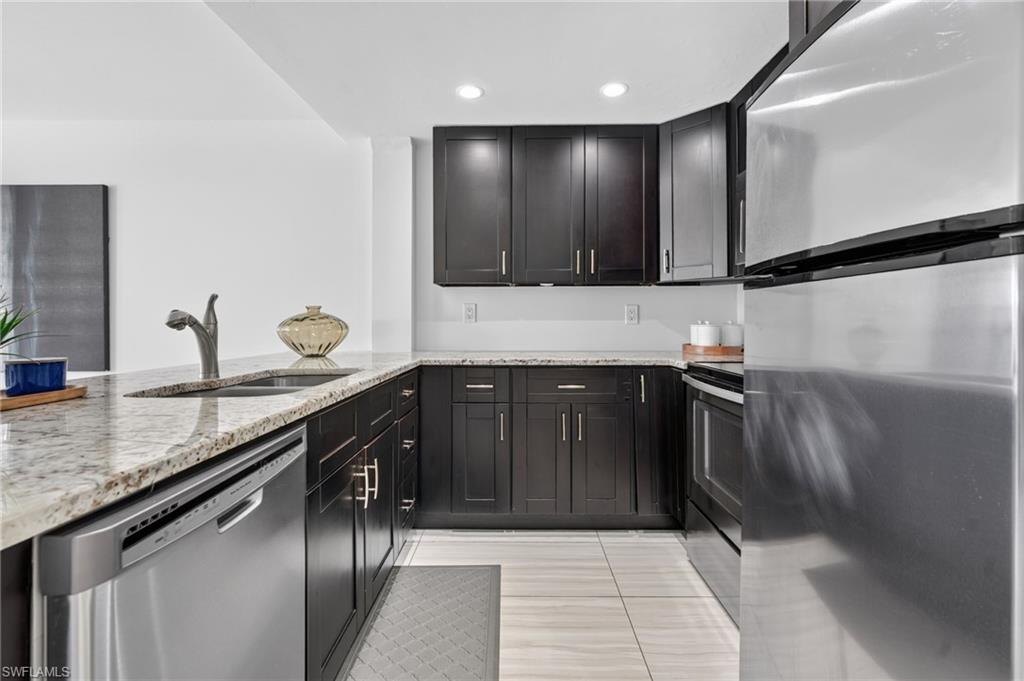 Galley kitchen featuring dark wood cabinetry, light granite countertops, stainless steel appliances, a built-in sink, and recessed lighting
