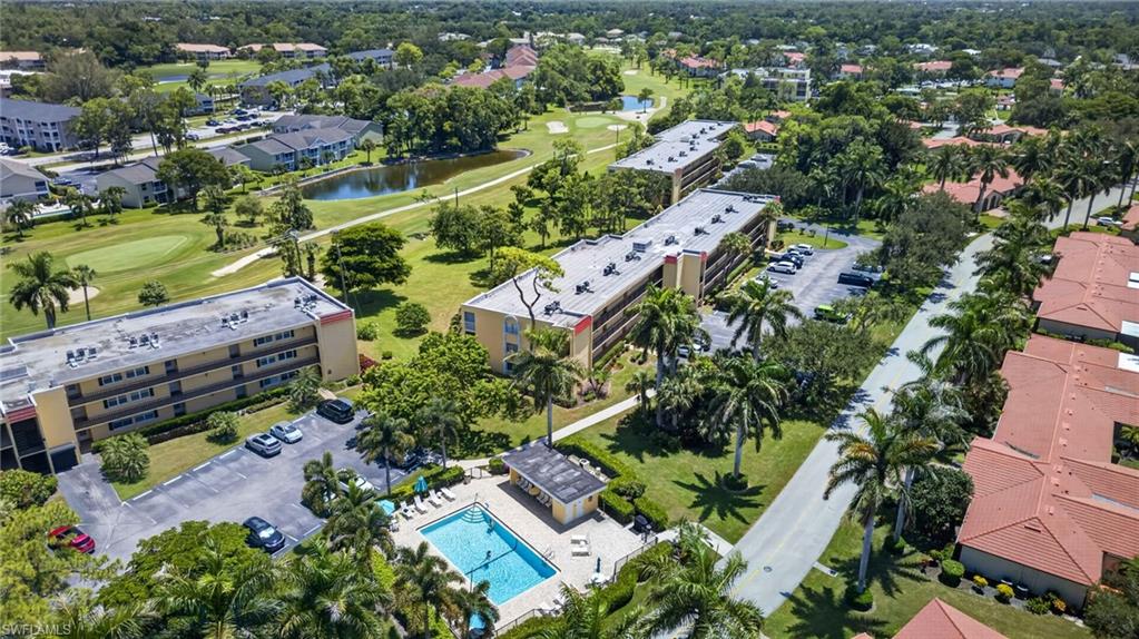 Aerial view showcasing a resort-style community with manicured grounds, mature palm and shade trees, resort-style swimming pool with surrounding hardscape, multiple residential buildings with modern architecture, and abundant green space throughout the property