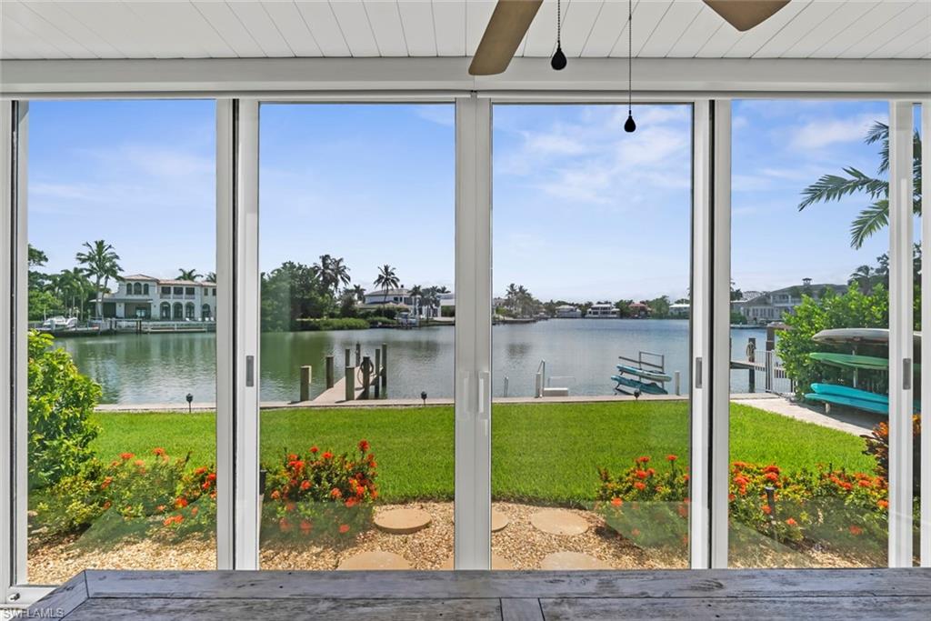 Expansive sliding glass doors opening to a waterfront backyard with a manicured lawn, a private dock with pilings, and lush tropical landscaping