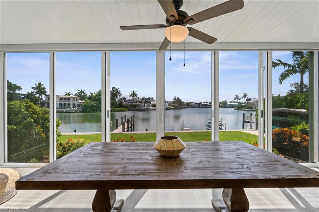 Expansive waterfront living space featuring floor-to-ceiling sliding glass doors, a large wooden dining table, a white beadboard ceiling, and a ceiling fan with light fixture