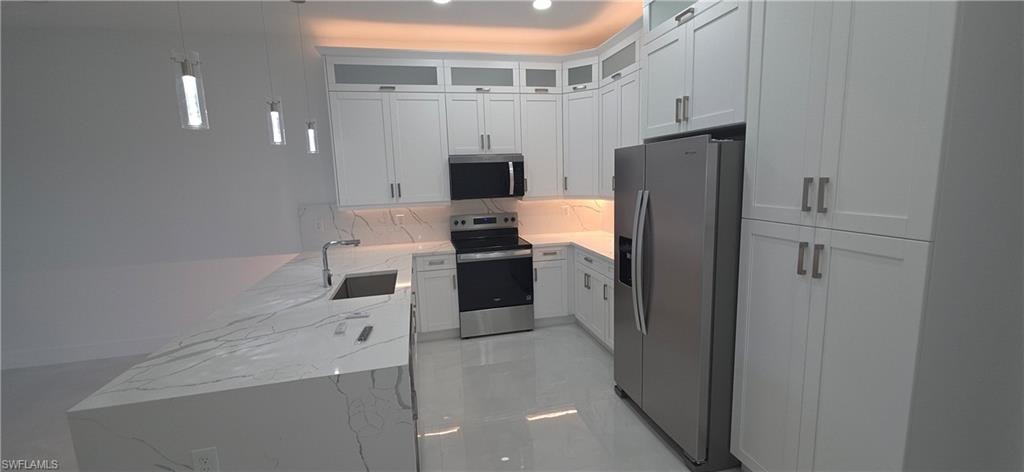 Modern kitchen featuring an expansive island with a waterfall-edge countertop and undermount sink, white shaker-style cabinetry, stainless steel appliances, and sleek pendant lighting