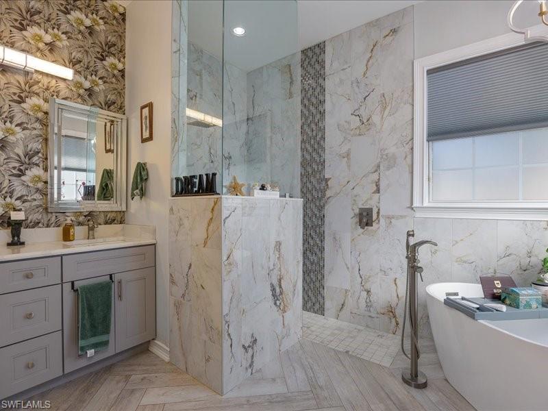 Contemporary bathroom featuring a freestanding soaking tub, a spacious walk-in shower with glass enclosure, a double vanity with light gray cabinetry, wall-mounted floral wallpaper, and large format tile flooring in a herringbone pattern