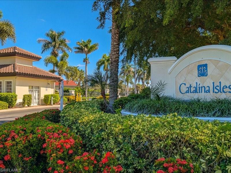 Gated community entrance featuring a sign with "Catalina Isles" branding, manicured landscaping with red flowering shrubs, and a guardhouse with a tiled roof