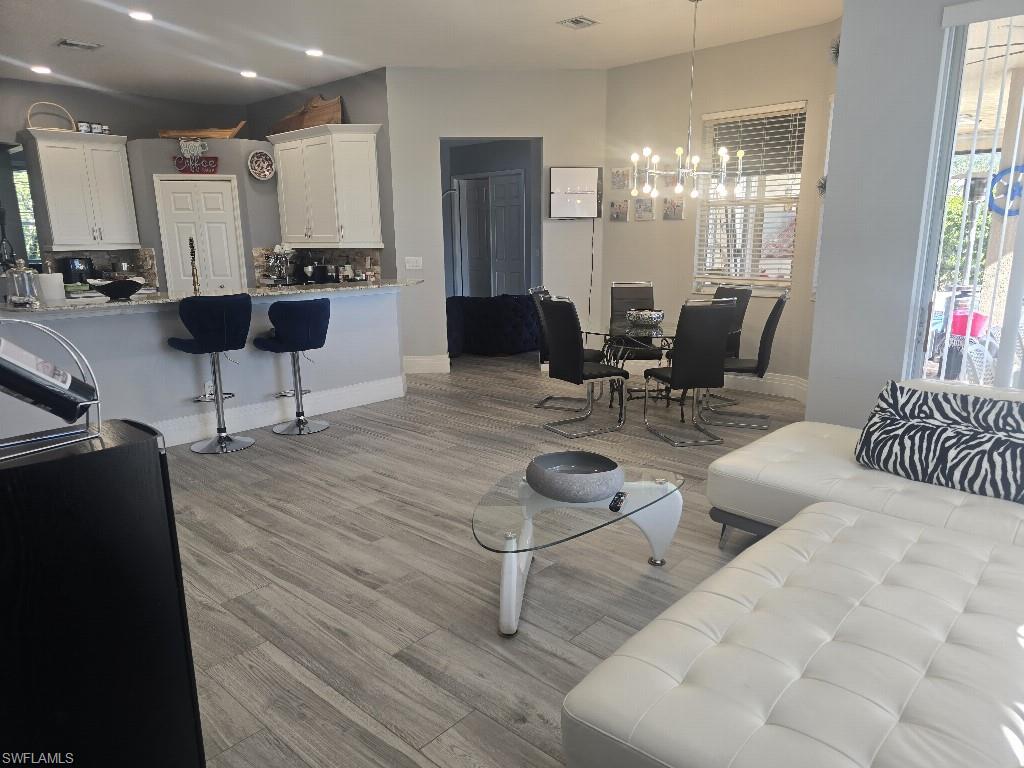 Open-concept living area featuring wood-look tile flooring, a kitchen island with seating, white shaker-style cabinetry, and a dining area with a modern chandelier