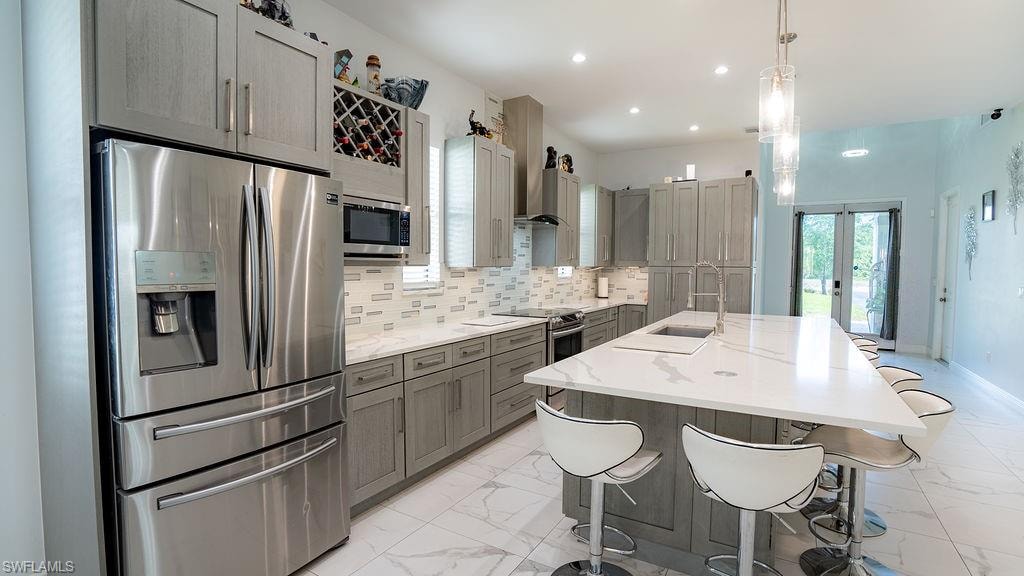 Modern kitchen featuring light gray cabinetry, stainless steel appliances, a large center island with a built-in sink, white marble-look countertops, and white tile flooring