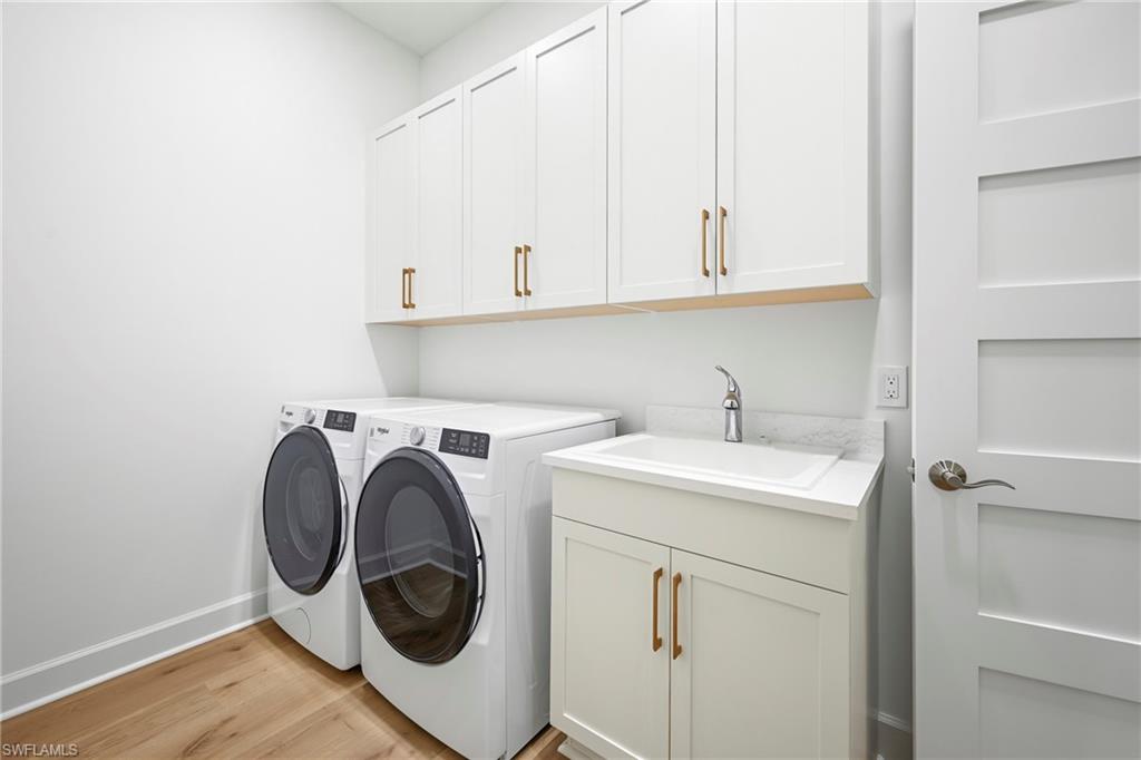 Dedicated laundry area featuring wood-finish flooring, white cabinetry with bronze hardware, a utility sink with a stone-finish countertop, and a contemporary four-panel interior door