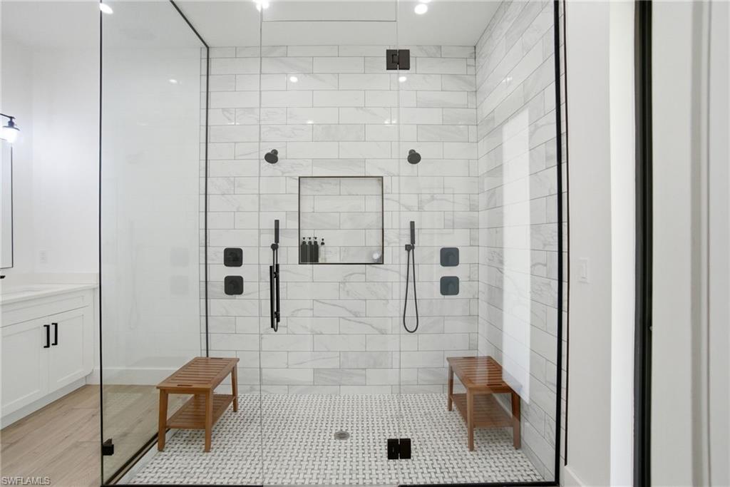 Spacious walk-in shower featuring a glass enclosure with matte black trim, white subway tile walls, a mosaic tile floor, dual shower heads, and an integrated shower niche