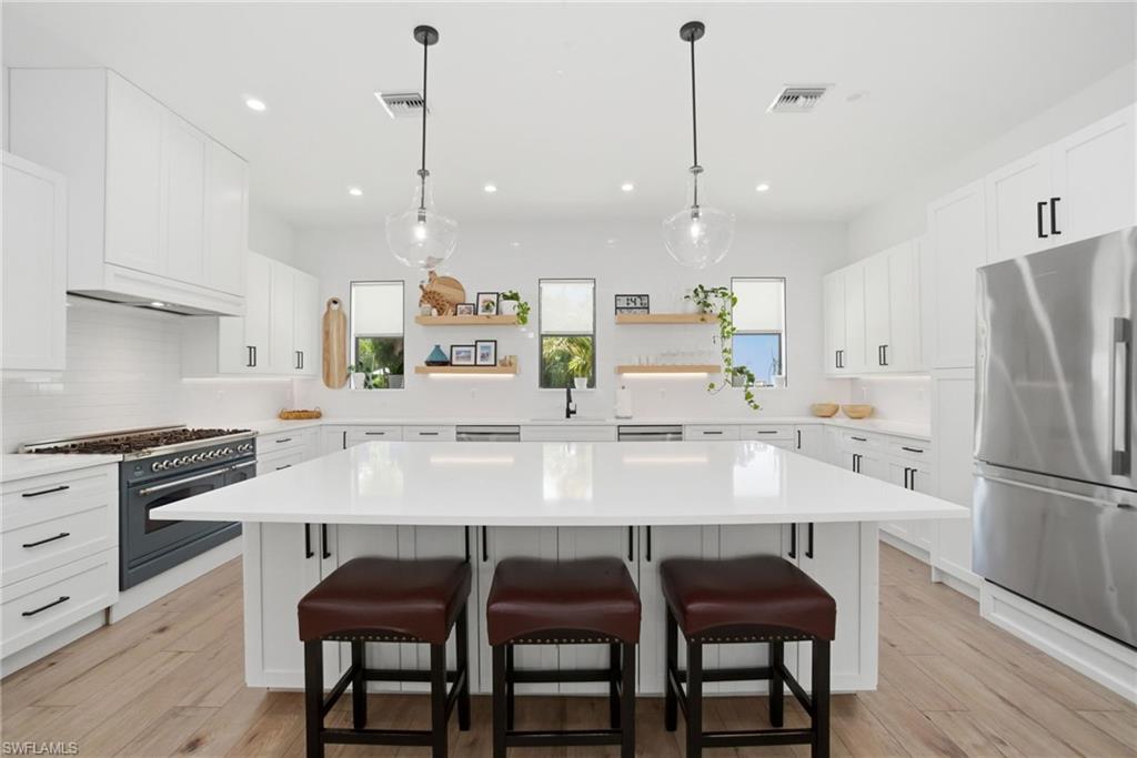 Spacious kitchen featuring an expansive central island, white cabinetry with black hardware, stainless steel appliances, wood-finish flooring, and pendant lighting