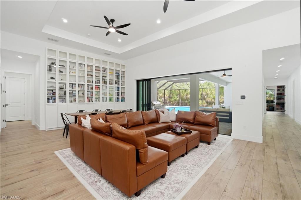 Spacious living area featuring light wood-finish flooring, a tray ceiling with recessed lighting and ceiling fans, and an expansive built-in white cabinetry wall with glass-fronted upper sections