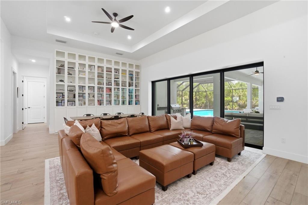 Spacious living area featuring light wood-finish flooring, a tray ceiling with recessed lighting and a ceiling fan, and extensive built-in shelving with glass-front cabinets