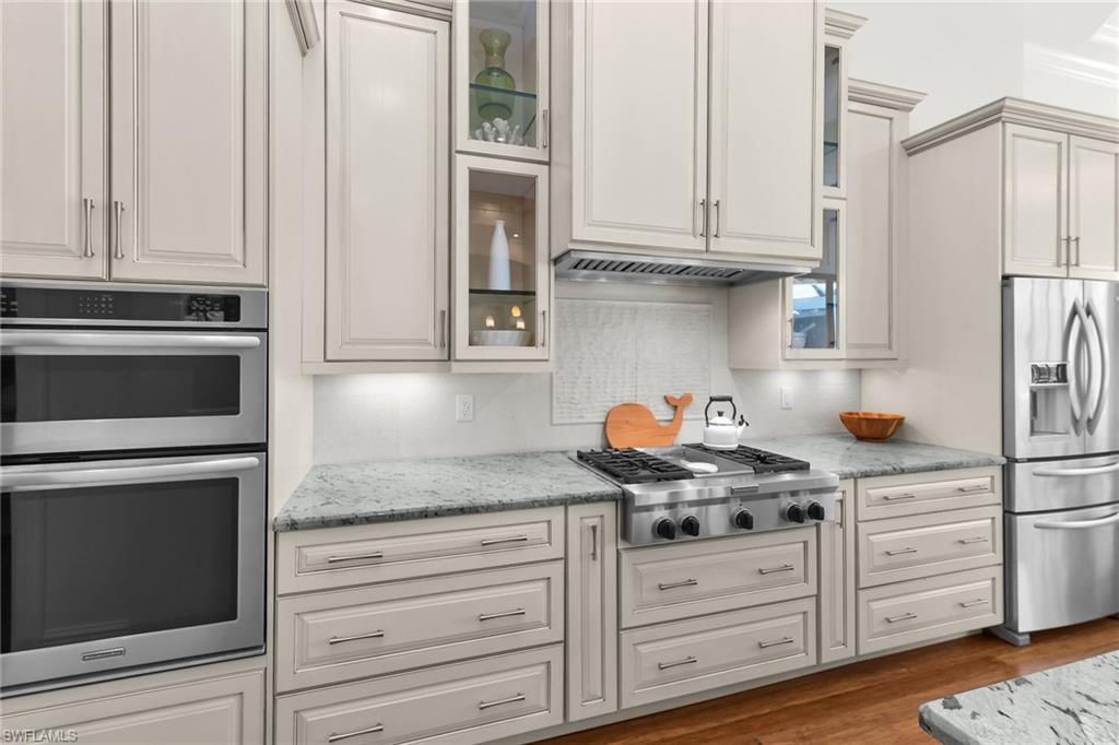 Kitchen featuring light-toned cabinetry, stainless steel appliances, a gas range with a matching range hood, a tiled backsplash, and stone-finish countertops