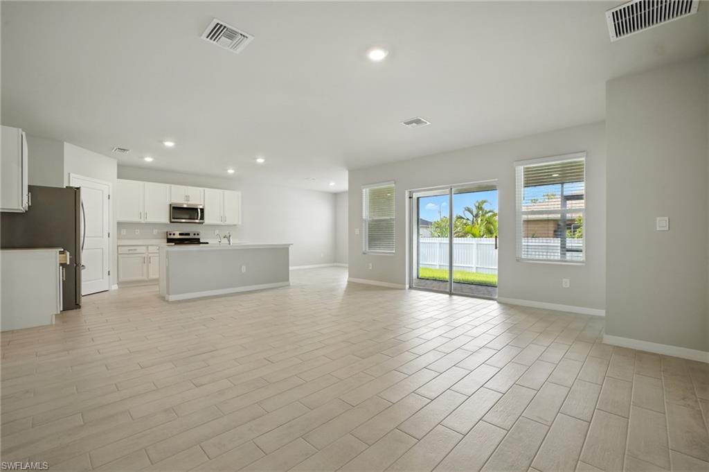 Spacious open-concept living area featuring light-toned tile flooring, recessed lighting, white kitchen cabinetry with stainless steel appliances, a kitchen island with a breakfast bar, and sliding glass doors to the exterior