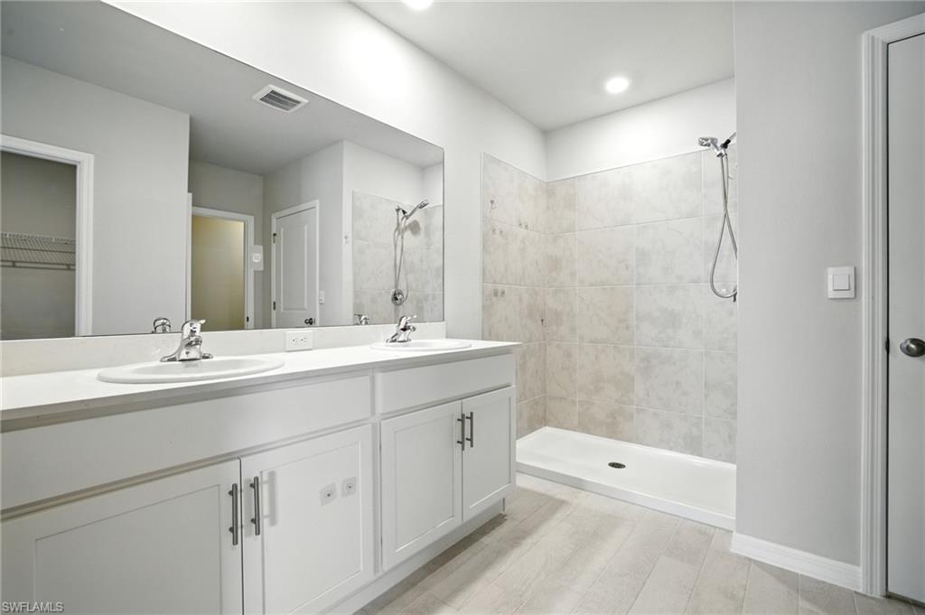 Bathroom featuring a double vanity with white cabinetry and chrome fixtures, a broad mirror, and a walk-in shower with tiled walls and a handheld showerhead