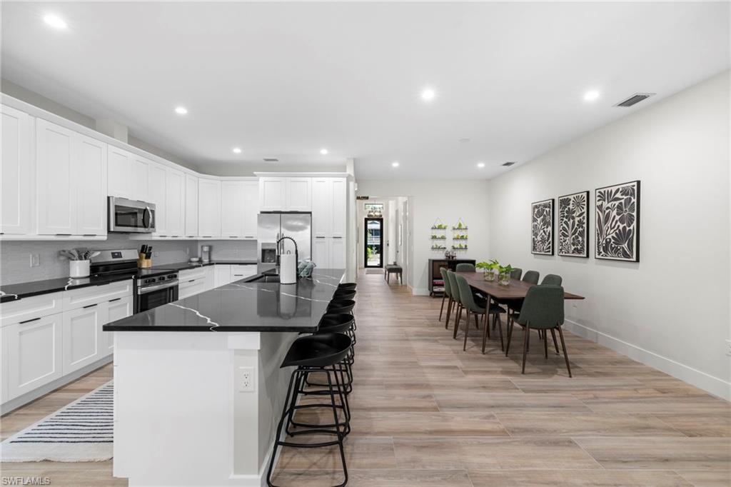 Spacious kitchen and dining area featuring an expansive island with seating, white shaker cabinetry, stainless steel appliances, dark countertops with light veining, and light wood-finish flooring