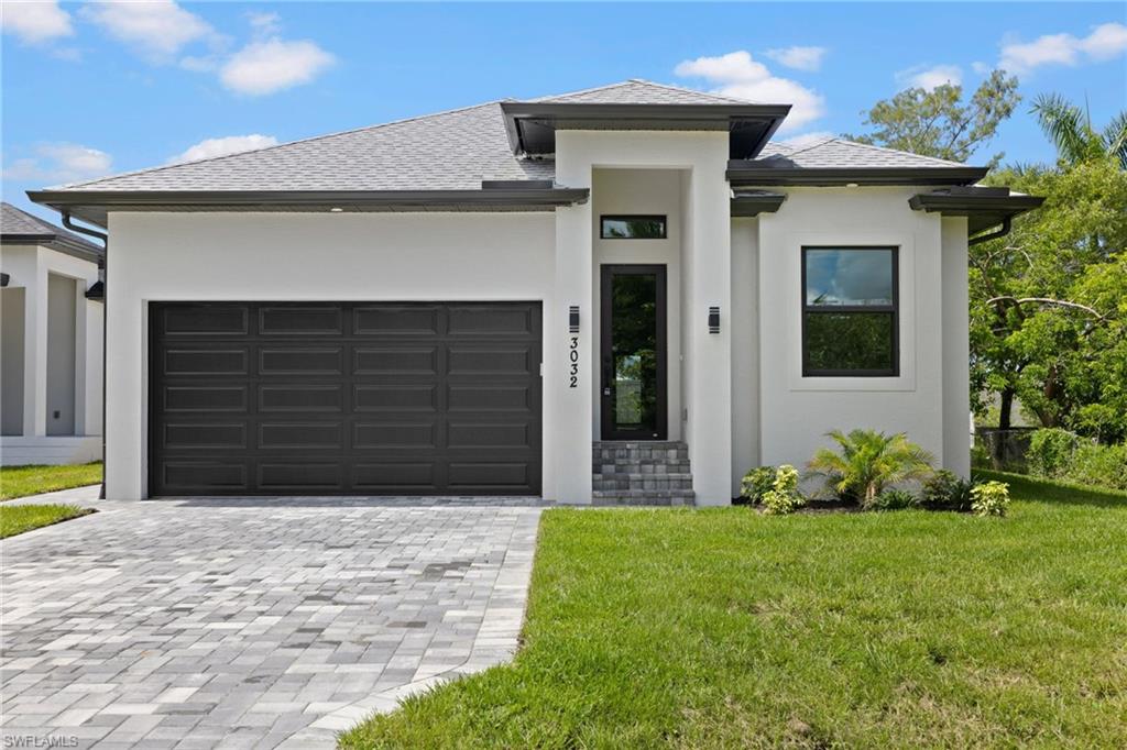 Contemporary stucco exterior featuring a dark paneled garage door, paver driveway, dark-framed windows, and decorative landscaping