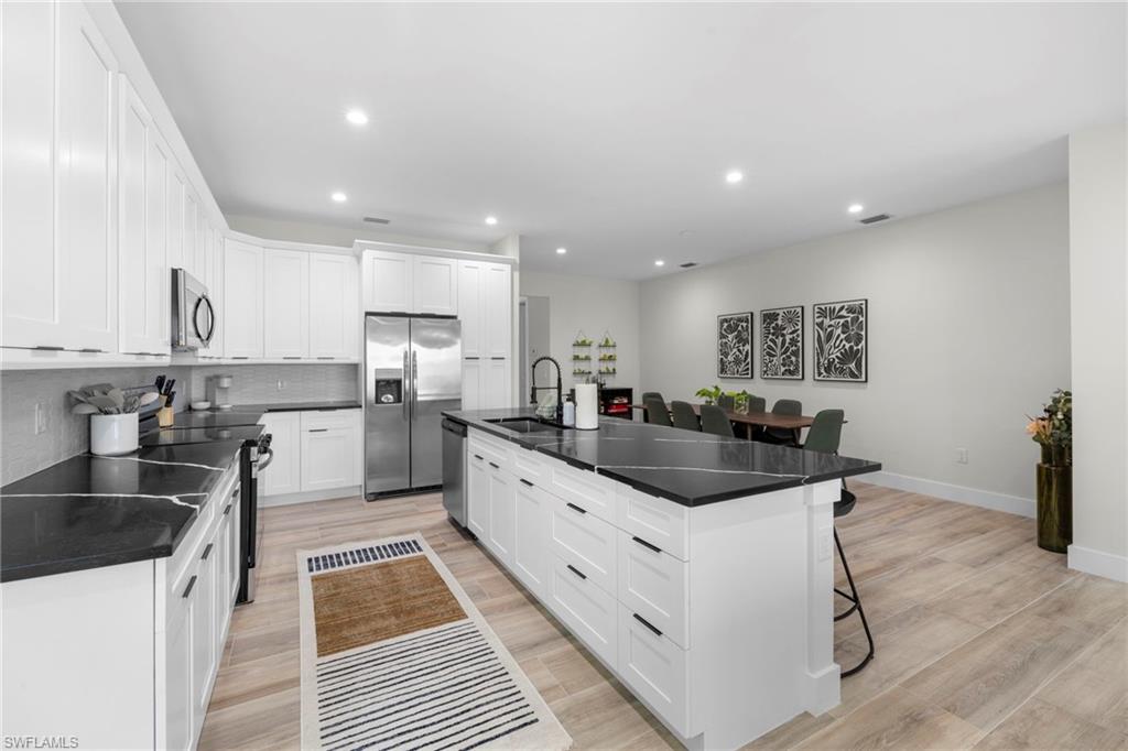 Modern kitchen featuring a large central island with seating, dark countertops, white shaker cabinetry, stainless steel appliances, and wood-finish flooring