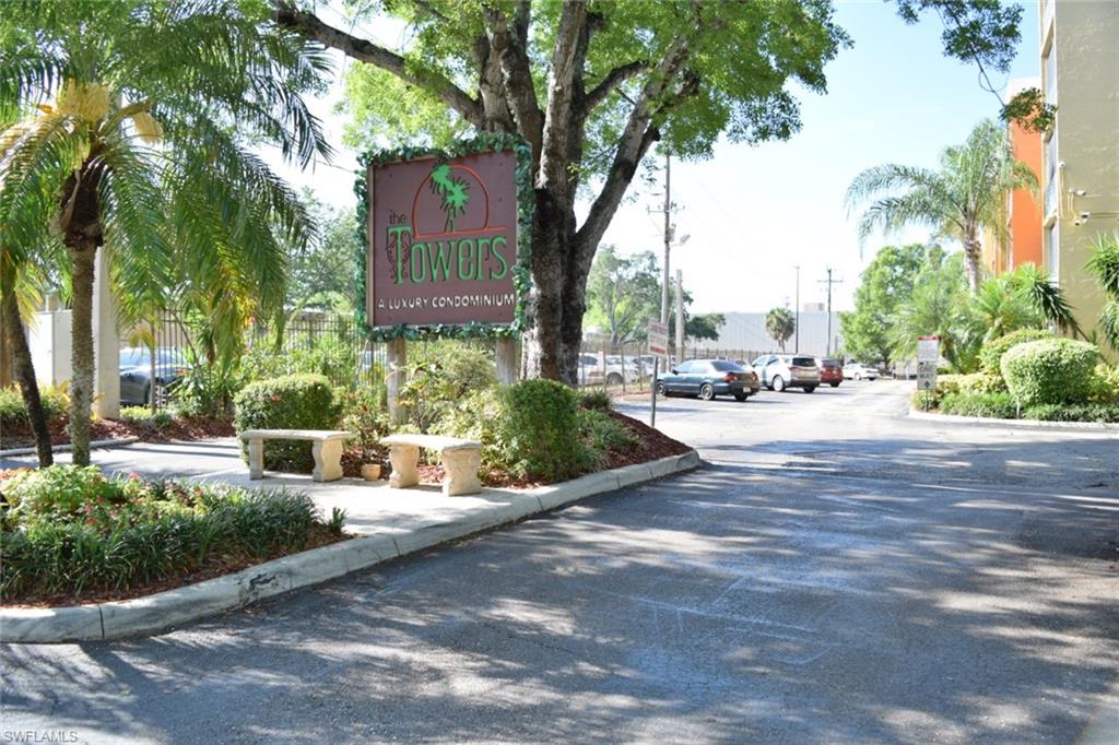 Entryway featuring a prominent community sign, stone benches, lush landscaping, mature trees, and palm trees