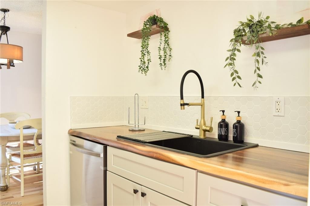 Kitchen featuring a black composite sink with a matte gold and black faucet, natural wood countertops, white shaker-style cabinetry with black hardware, white hexagonal tile backsplash, and a stainless steel dishwasher
