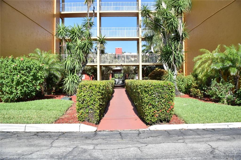 Exterior building facade with multiple balconies, featuring a red walkway bordered by manicured hedges and lush landscaping