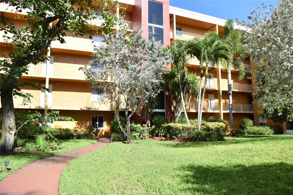 Multi-story building featuring an exterior elevator, private balconies with white railings, and lush landscaping including mature trees and palm trees