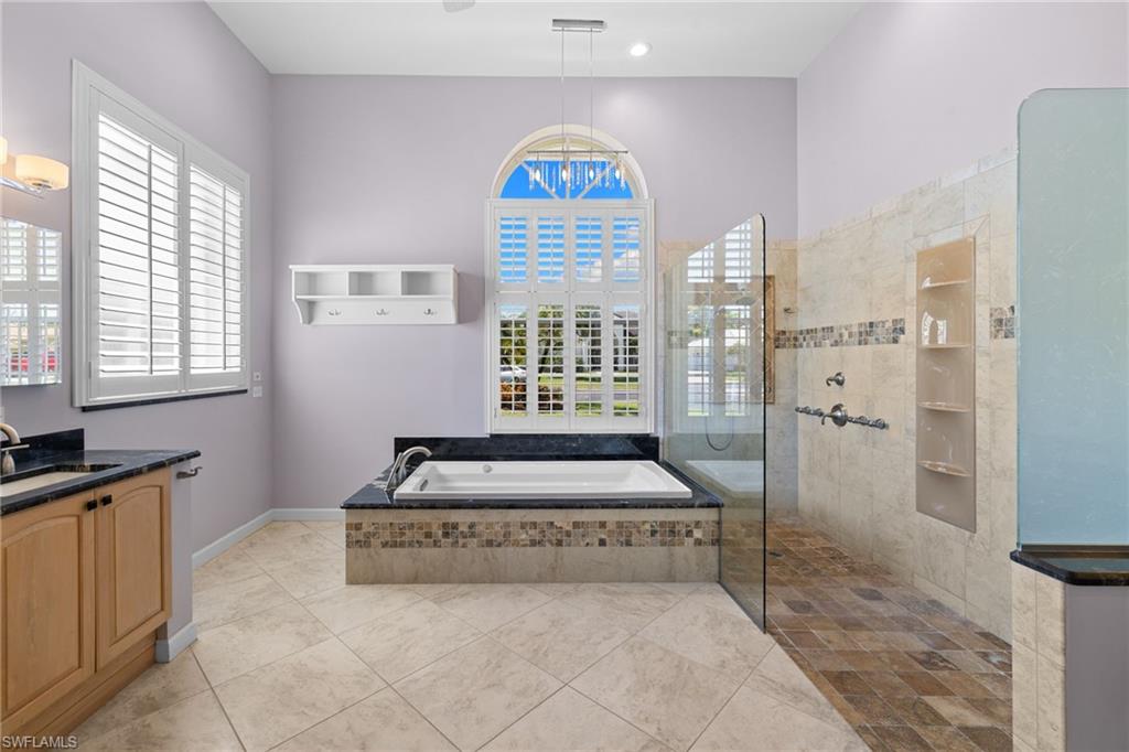 Primary bathroom #1 featuring a built-in soaking tub with mosaic tile surround, a separate walk-in shower with glass enclosure and tiled niche, dual vanity with black countertops, and plantation-shuttered windows