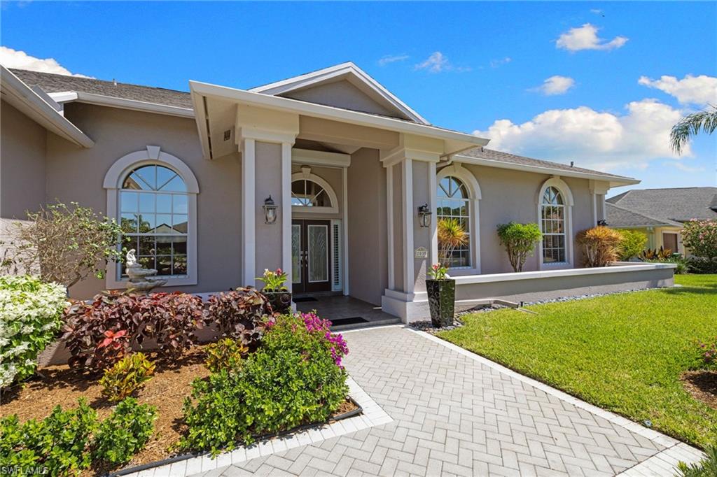 Exterior entry featuring a covered portico with substantial columns, arched windows, a double front door, a paver walkway, and mature landscaping