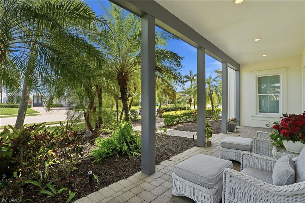 Covered porch featuring a textured paver floor, recessed lighting, substantial support columns, and an integrated garden bed with mature palm trees and diverse landscaping