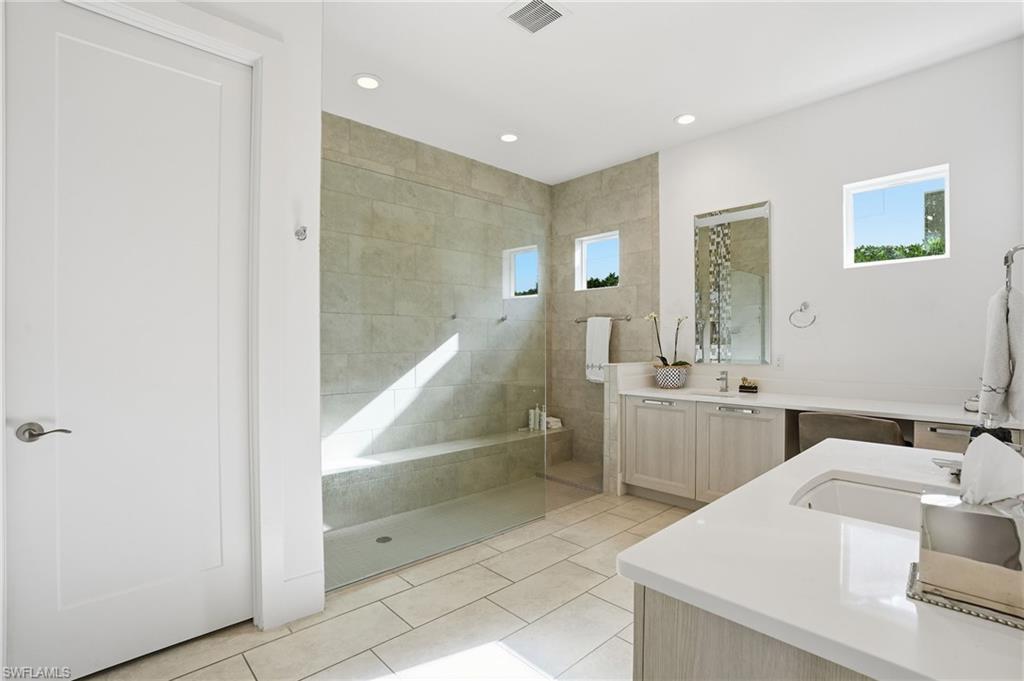 Spacious bathroom featuring a glass-enclosed shower with tiled walls and a built-in bench, dual vanity with light-toned cabinetry and white countertops, and recessed lighting